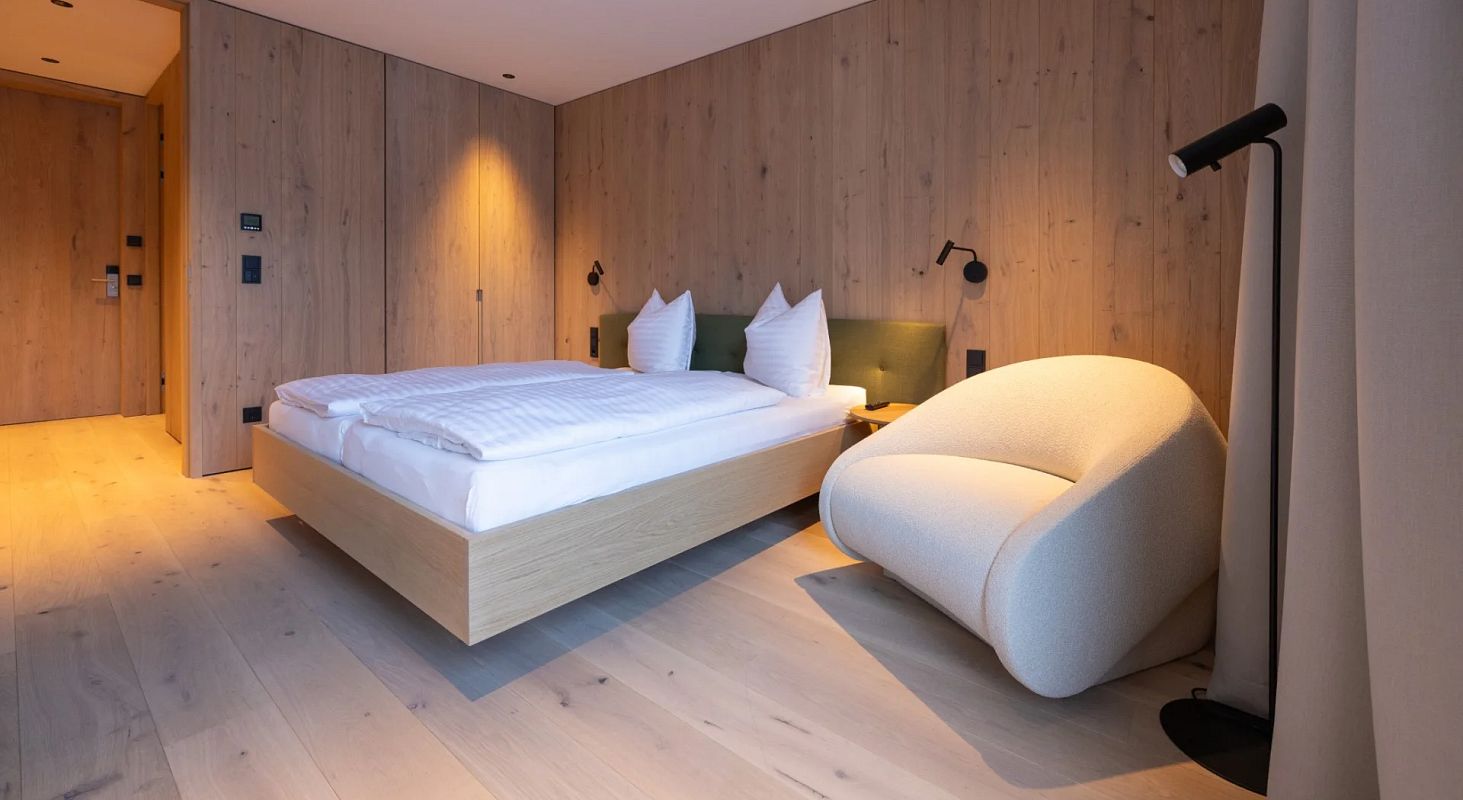 Modernes Schlafzimmer mit hellem Holzboden und Wänden, einem großen Bett mit weißen Kissen und Bettzeug, sowie einem stilvollen, weißen Sessel daneben.