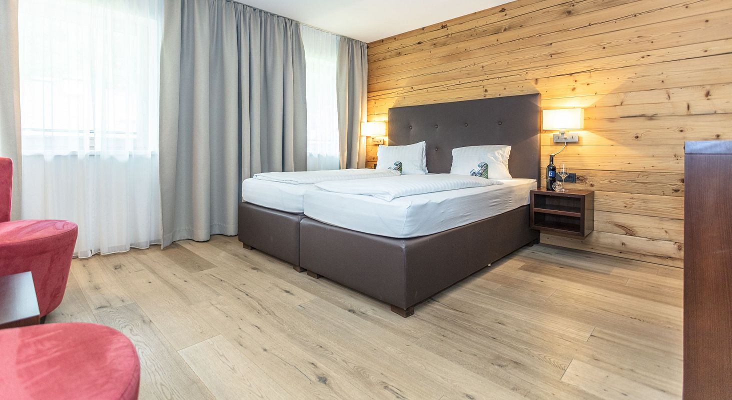 Gemütliches Hotelzimmer mit Holzwänden, zwei zusammengestellten Einzelbetten, großen Vorhängen, einem roten Sessel und warmer Beleuchtung.