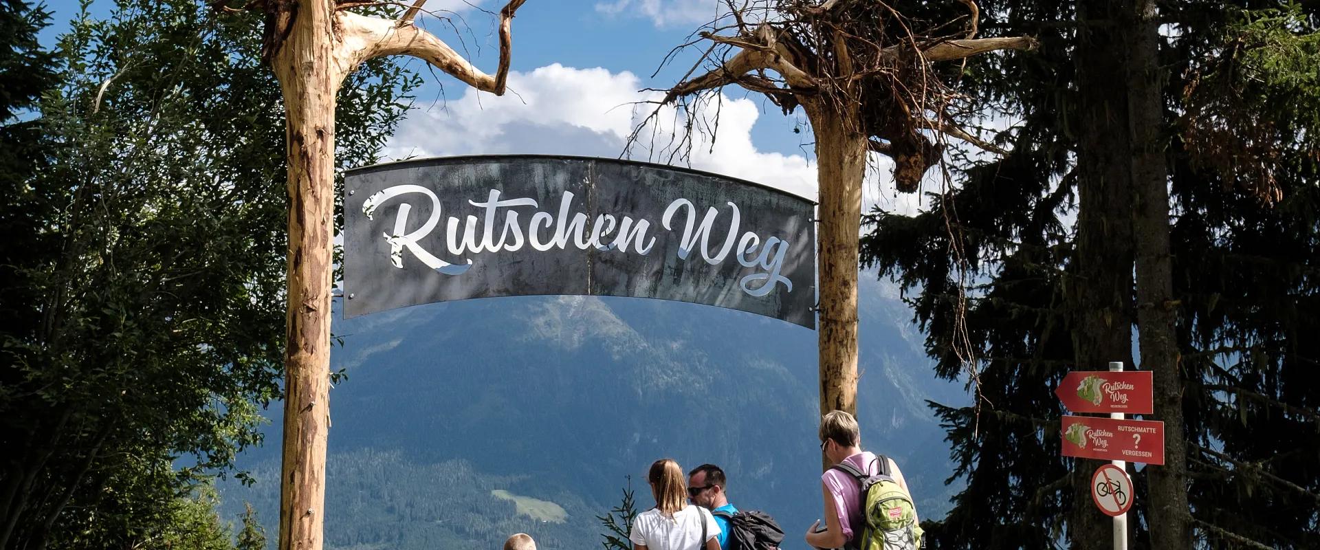 Eine Familie steht vor einem Holzportal mit der Aufschrift "Rutschen Weg" in einer bergigen Landschaft mit Bäumen und einem klaren Himmel.