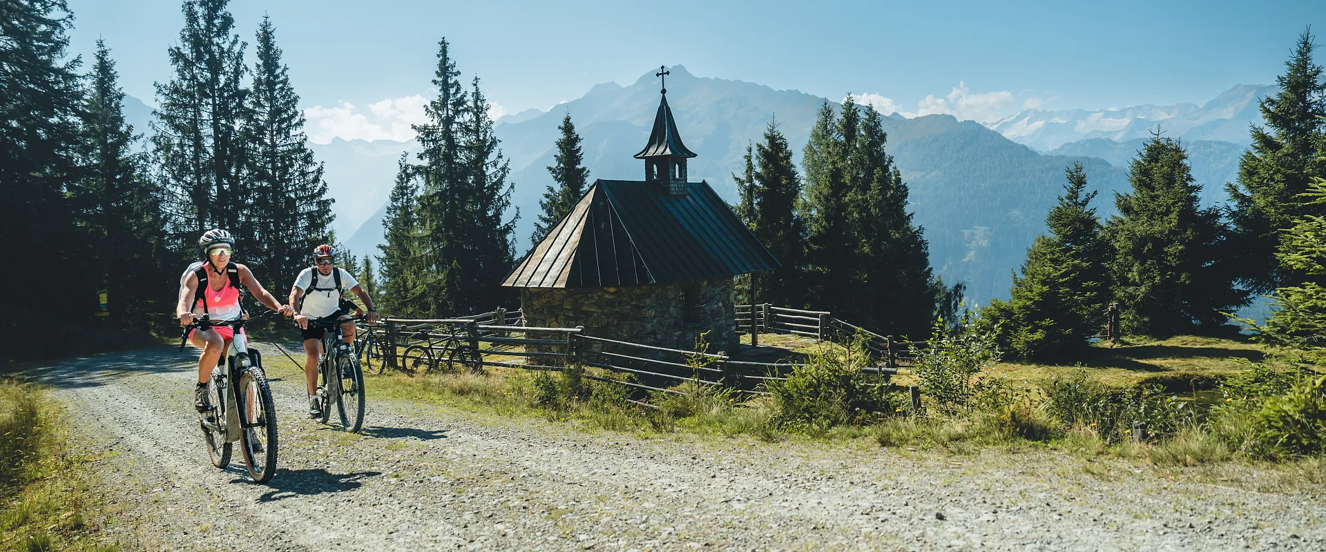 Zwei Radfahrer fahren auf einem Schotterweg durch eine alpine Landschaft mit einer kleinen Kapelle im Hintergrund, umgeben von Bäumen und Bergen bei klarem Himmel.