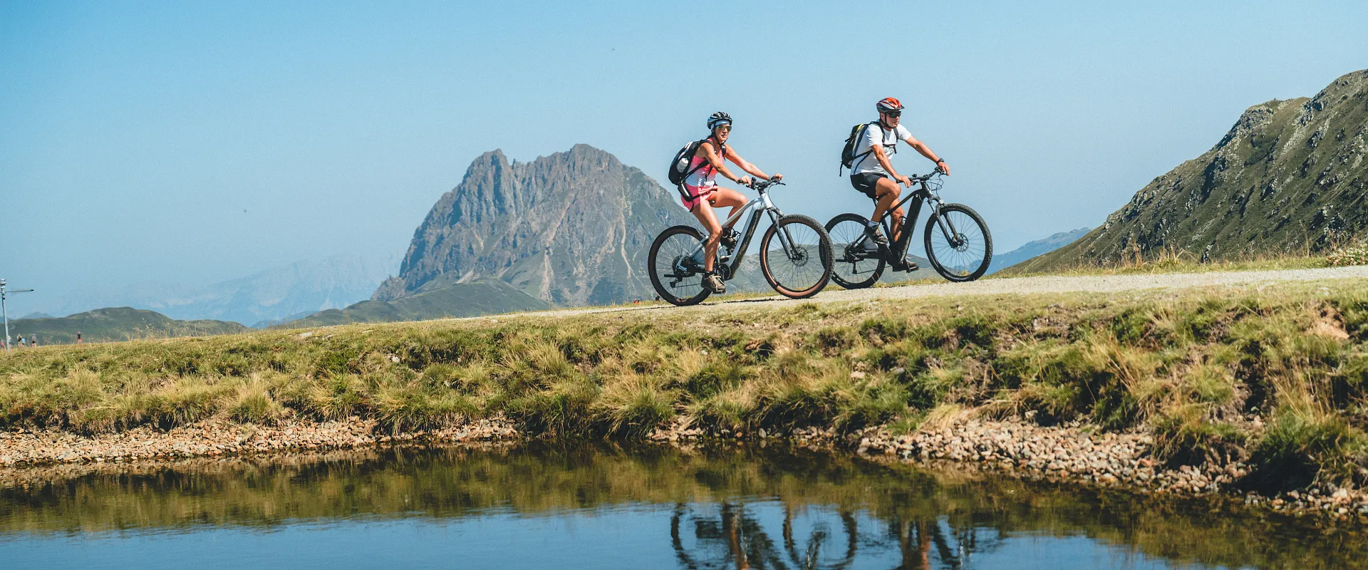 Zwei Personen fahren mit Mountainbikes auf einem Pfad entlang eines klaren Sees. Im Hintergrund sind grüne Hügel und ein markanter Berg zu sehen.