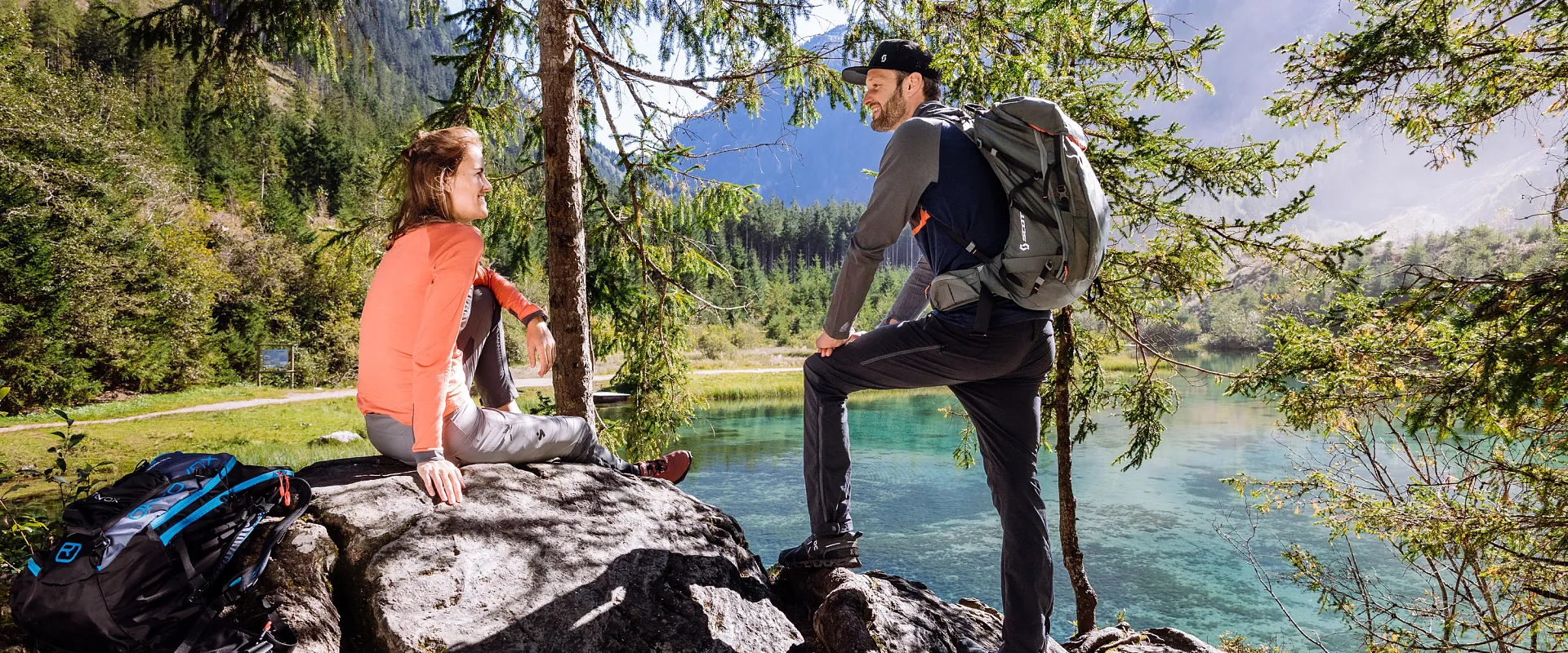 Zwei Personen sitzen auf einem Felsen am Ufer eines klaren Sees, umgeben von Bergen und Nadelwäldern, genießen die Natur und Sonnenschein.