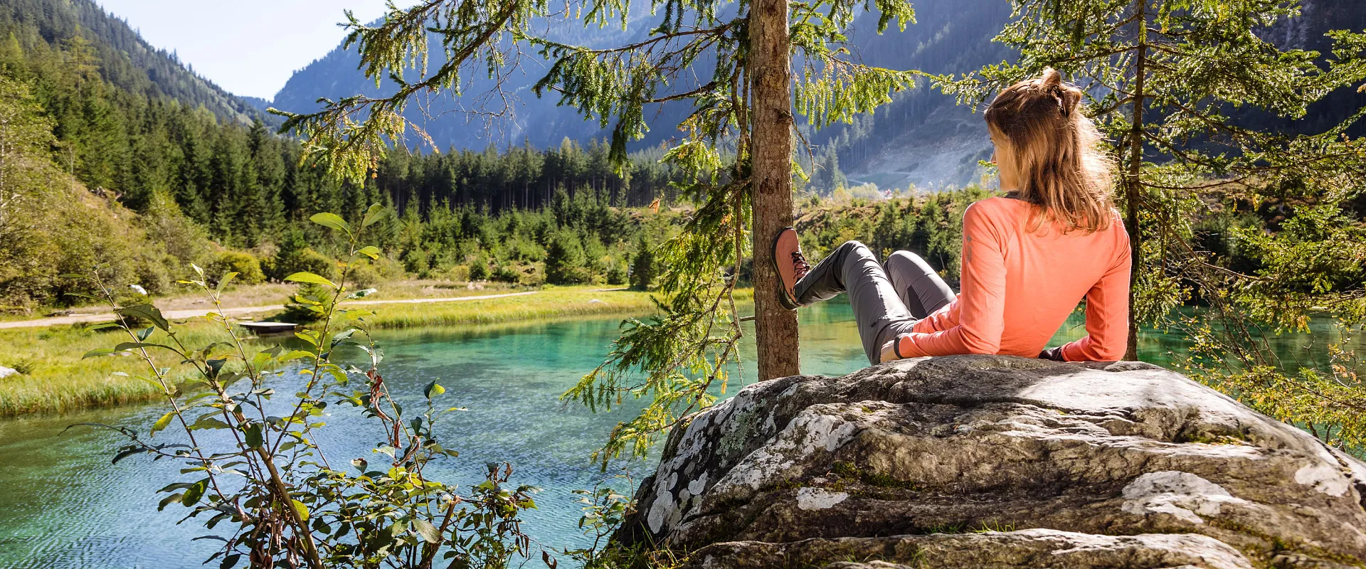 Eine Frau sitzt entspannt auf einem Felsen am Ufer eines klaren Sees, umgeben von hohen Bergen und grünem Wald, bei sonnigem Wetter.