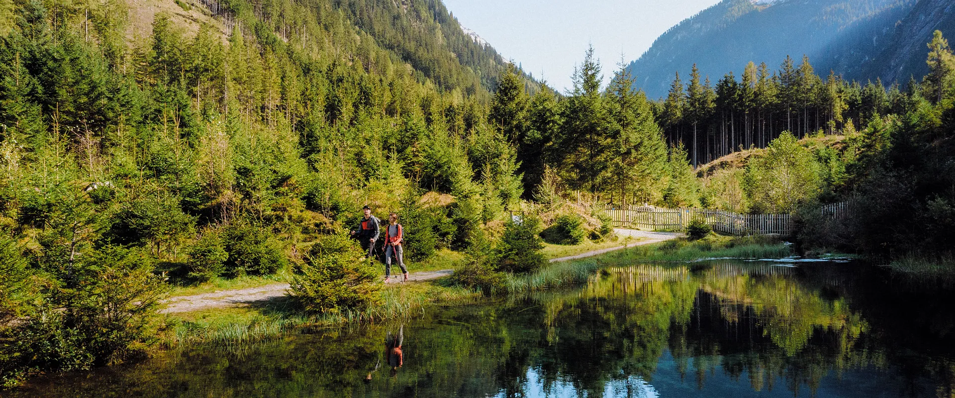 Ein kleiner, klarer See inmitten grüner Nadelwälder, umgeben von hohen Bergen mit schneebedeckten Gipfeln unter einem klaren blauen Himmel.