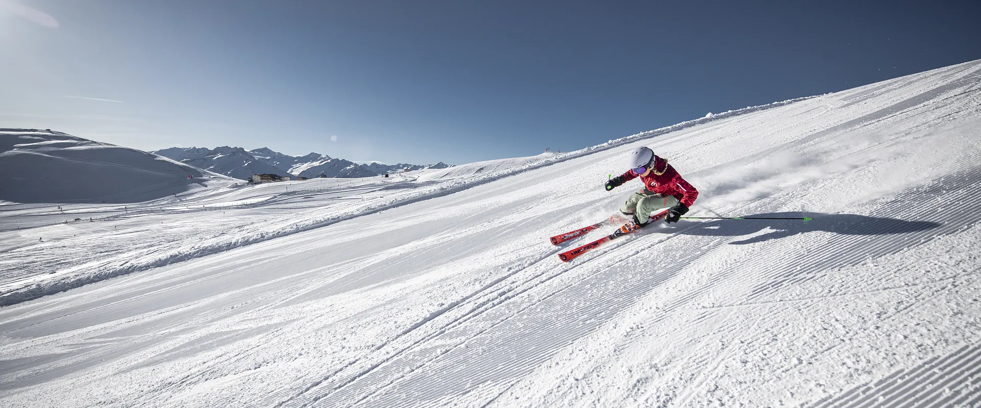 Eine Person in roter Skiausrüstung fährt dynamisch auf einer schneebedeckten Piste unter klarem, blauem Himmel. Im Hintergrund sind schneebedeckte Berge zu sehen.