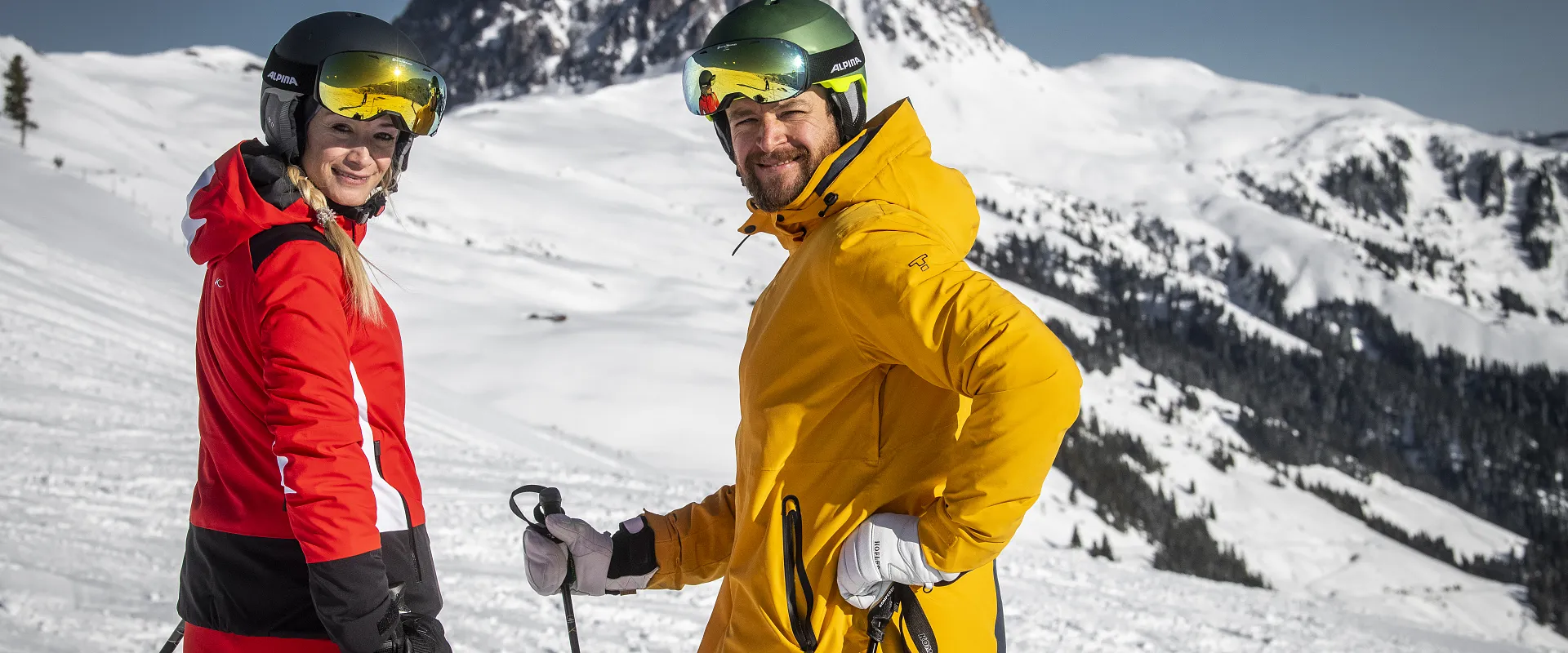 Zwei Personen in leuchtend roten und gelben Skianzügen stehen mit Skiern auf einer verschneiten Berglandschaft. Im Hintergrund ein hoher, schneebedeckter Berg.