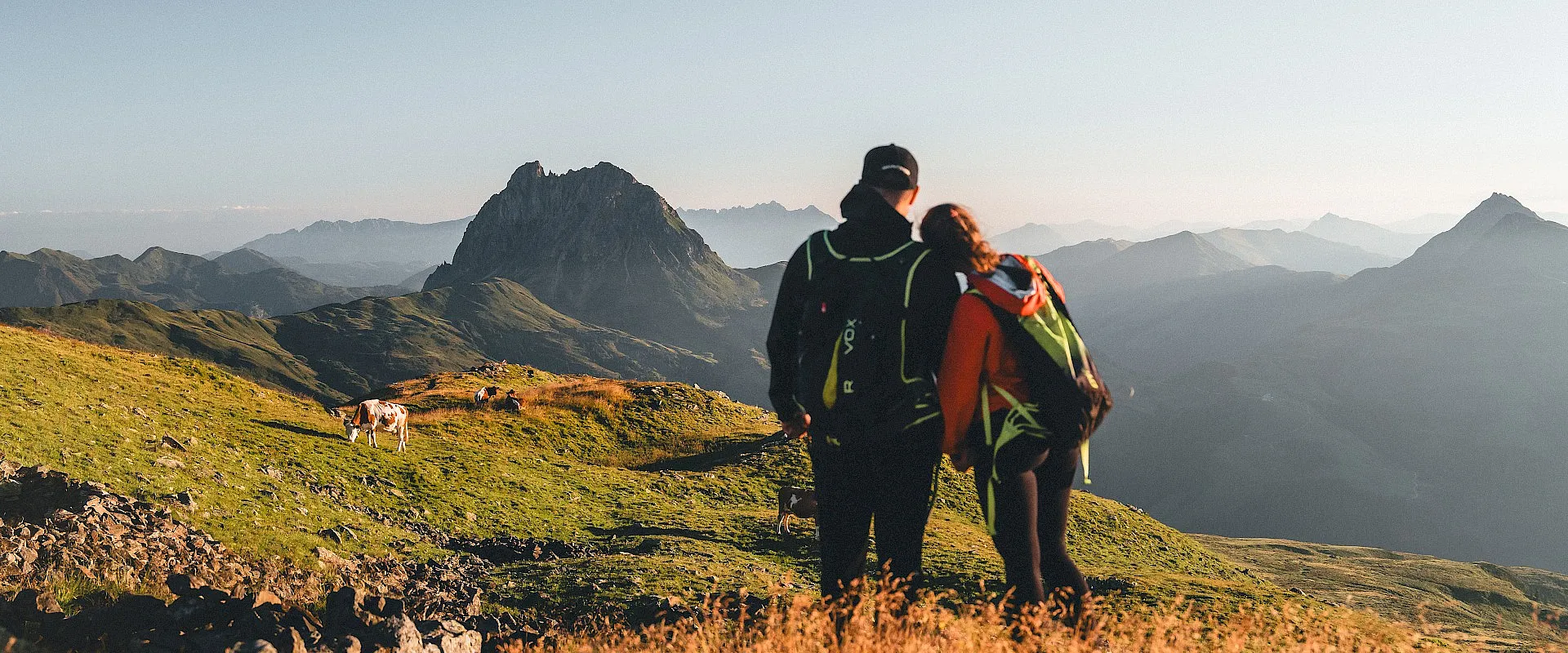 Paar auf einer Bergwanderung, steht in einer grünen Landschaft mit Bergen im Hintergrund, bei Sonnenuntergang. Sie tragen Rucksäcke und Outdoor-Bekleidung.