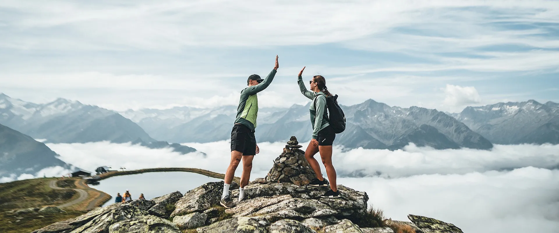 Zwei Menschen geben sich ein High-Five auf einem Berggipfel, umgeben von atemberaubender Berglandschaft und Wolken, die die Täler darunter bedecken.