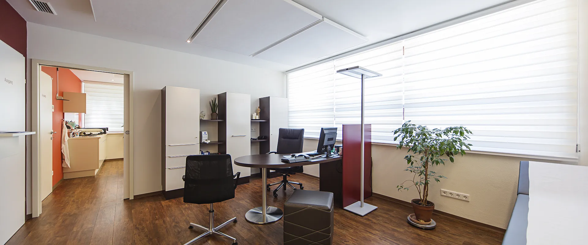 Modernes Büro mit Holzboden, weißen Wänden und einem großen Fenster. Ein Schreibtisch mit zwei Bürostühlen steht zentral, daneben ein kleiner Tisch mit Pflanzen.