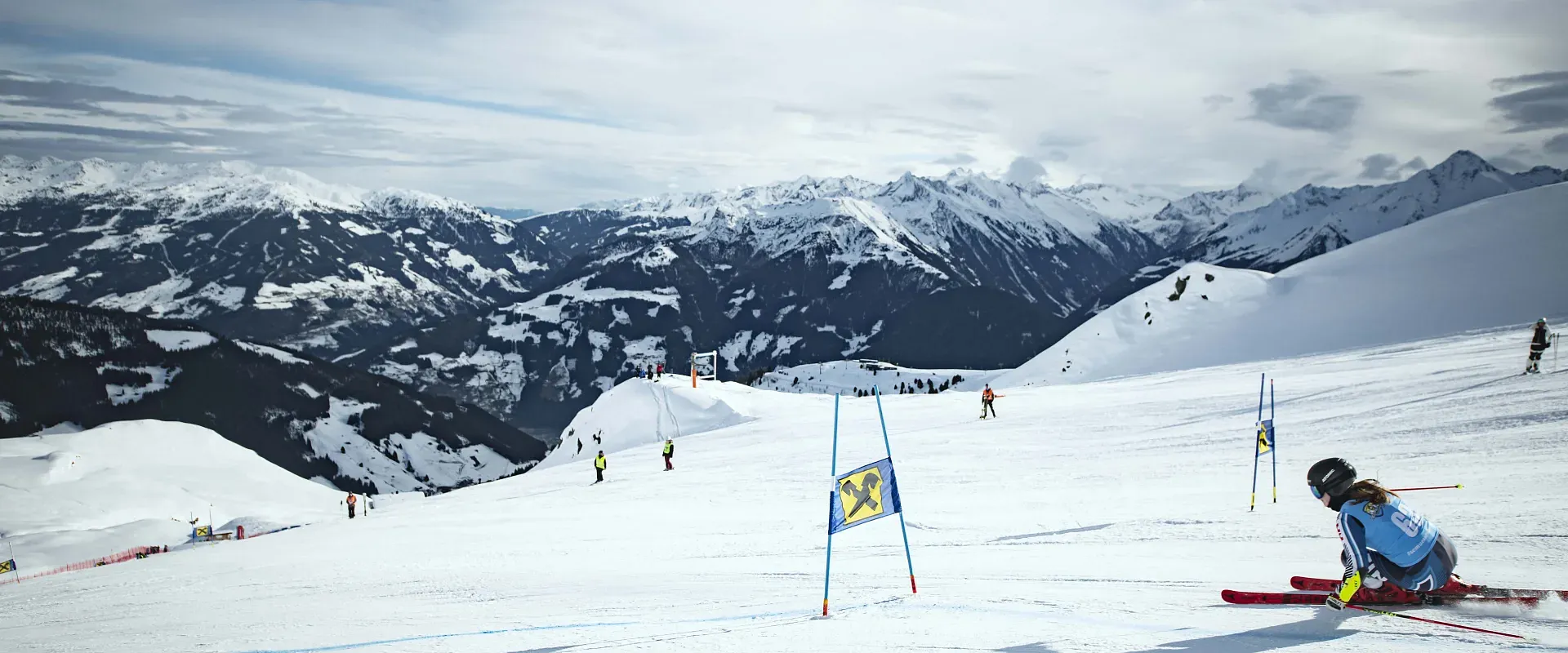 Ein Skifahrer in voller Fahrt auf einer verschneiten Skipiste in den Bergen. Der Himmel ist bewölkt und im Hintergrund sind majestätische Gebirgsketten zu sehen.