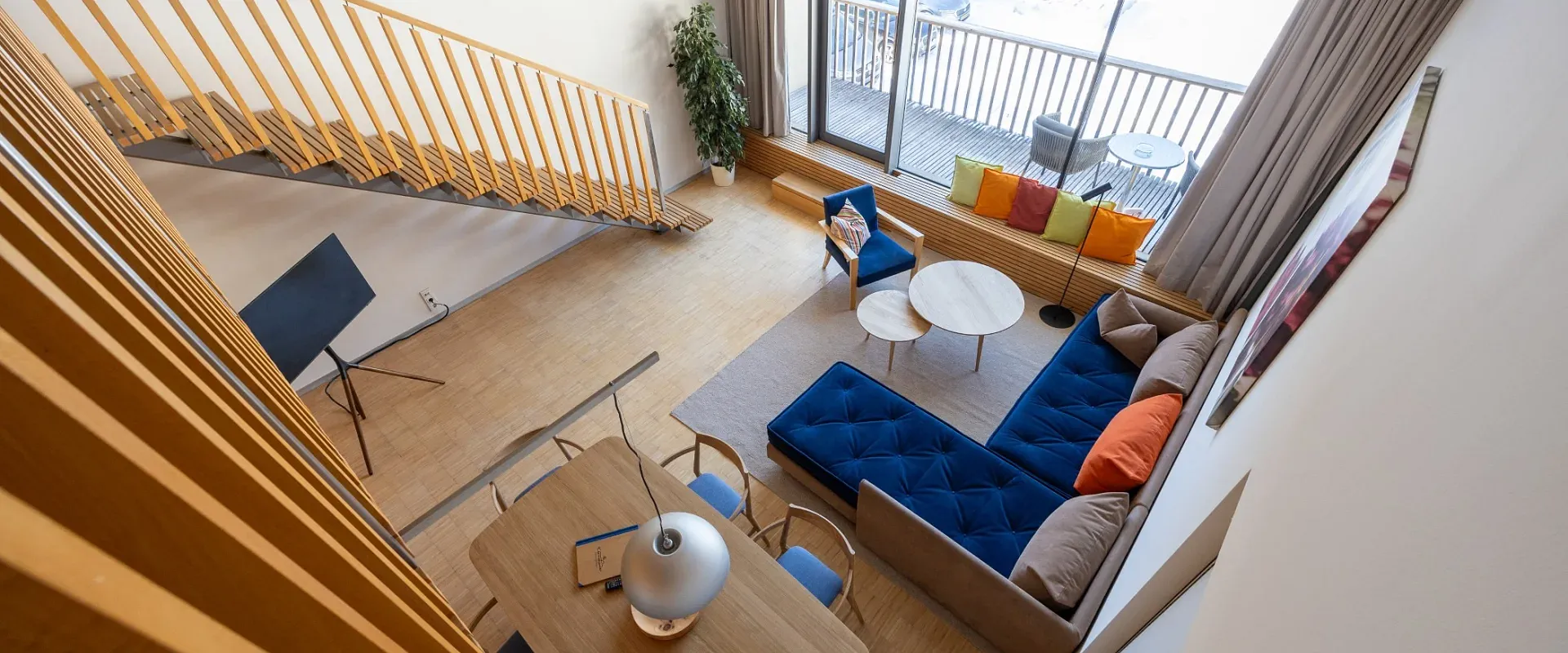 Moderne, minimalistische Wohnzimmereinrichtung mit blauer Couch, Holztisch, Stuhl und großen Fenstern. Helle Farben und viel natürliches Licht.