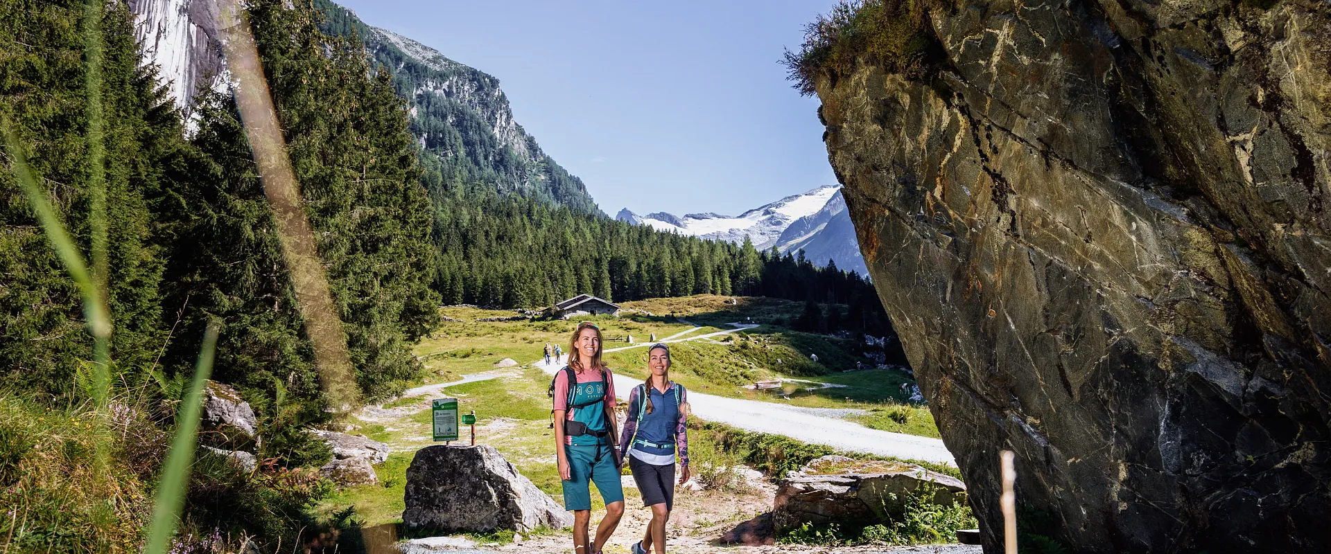 Zwei Personen wandern auf einem Pfad in einer Berglandschaft mit üppigem Grün, hohen Bäumen und fernen schneebedeckten Gipfeln unter blauem Himmel.