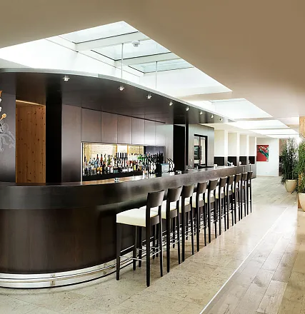 Moderne Hotelbar mit langer, geschwungener Theke, hohen Stühlen und eleganten Beleuchtung. An der Wand sind Regale mit Flaschen und Gläsern.