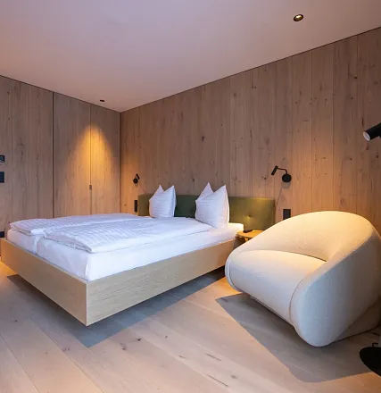 Modern eingerichtetes Schlafzimmer mit einem Doppelbett, Holzverkleidung an den Wänden und einem bequemen Sessel. Dezente Beleuchtung schafft eine gemütliche Atmosphäre.
