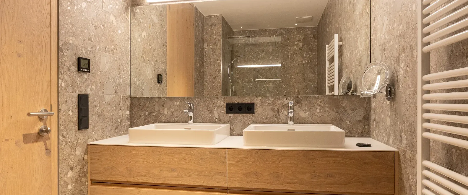 Modernes Badezimmer mit zwei Waschbecken auf einem Holzunterschrank, großen Spiegeln und dezenter Beleuchtung. Grau-marmorierte Wände und minimalistisches Design.