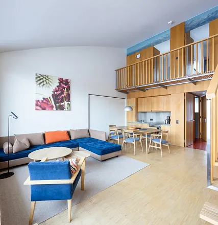 Modernes Wohnzimmer mit hohen Decken, einer blauen Sofagarnitur, Holzmöbeln und einer kleinen Küche. Eine Treppe führt zu einem offenen Zwischenboden.