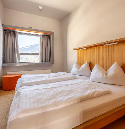 Ein helles, minimalistisches Hotelzimmer mit einem Doppelbett, weißen Bettdecken, einem Fenster mit Blick auf eine schneebedeckte Landschaft und einem orange Hocker.