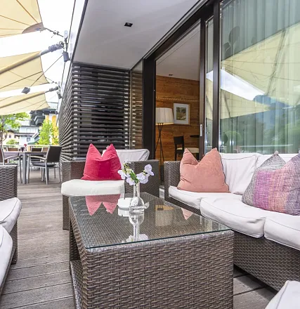 Moderne Terrasse mit bequemen Korbmöbeln und bunten Kissen. Ein Glastisch in der Mitte, umgeben von Pflanzen und großen Sonnenschirmen im Hintergrund.