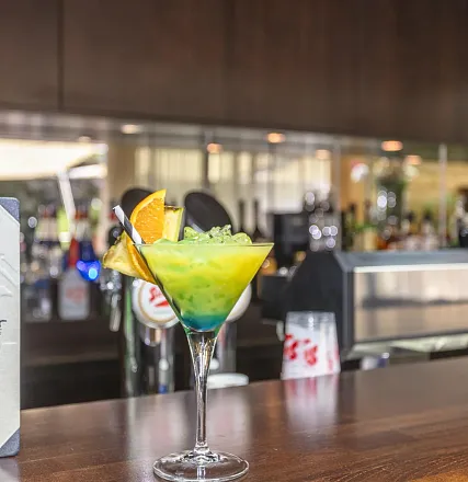 Ein farbenfroher Cocktail in einem eleganten Glas, geschmückt mit Orangenscheiben und einem Strohhalm, steht auf einer modernen Bar mit unscharfem Hintergrund.