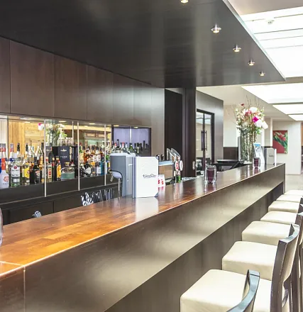 Elegante Bar mit einer langen, hölzernen Theke. Dahinter stehen Regale voller Flaschen. Man sieht Blumen und stilvolle Barhocker im modernen Ambiente.