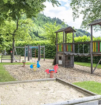 Ein Spielplatz mit Schaukeln, Wipptieren und einer Holzkletterstruktur. Umgeben von Bäumen und grünen Büschen, sonniges Wetter mit blauem Himmel.