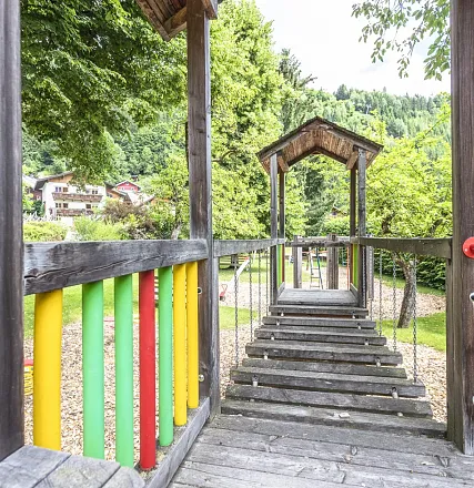 Ein bunter Spielplatz mit einer Hängebrücke aus Holz, umgeben von grünen Bäumen. Im Hintergrund sind Häuser und ein bewaldeter Hügel zu sehen.