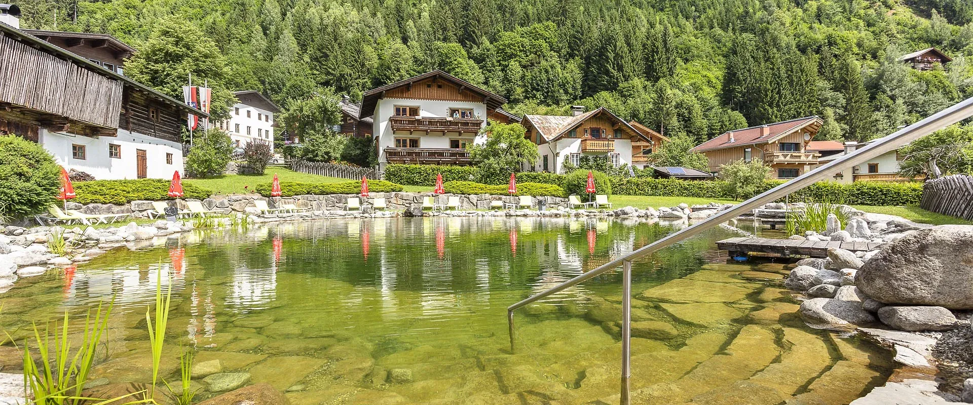 Landschaft mit natürlichem Schwimmteich, umrahmt von Steinen und Pflanzen. Im Hintergrund stehen traditionelle Holzchalets vor einer bewaldeten Hügelkulisse.
