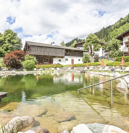 Ein malerischer Bergteich mit klarem Wasser, umgeben von Holzhäusern und grünen Bäumen. Rote Sonnenschirme stehen entlang des Ufers bei blauem Himmel.