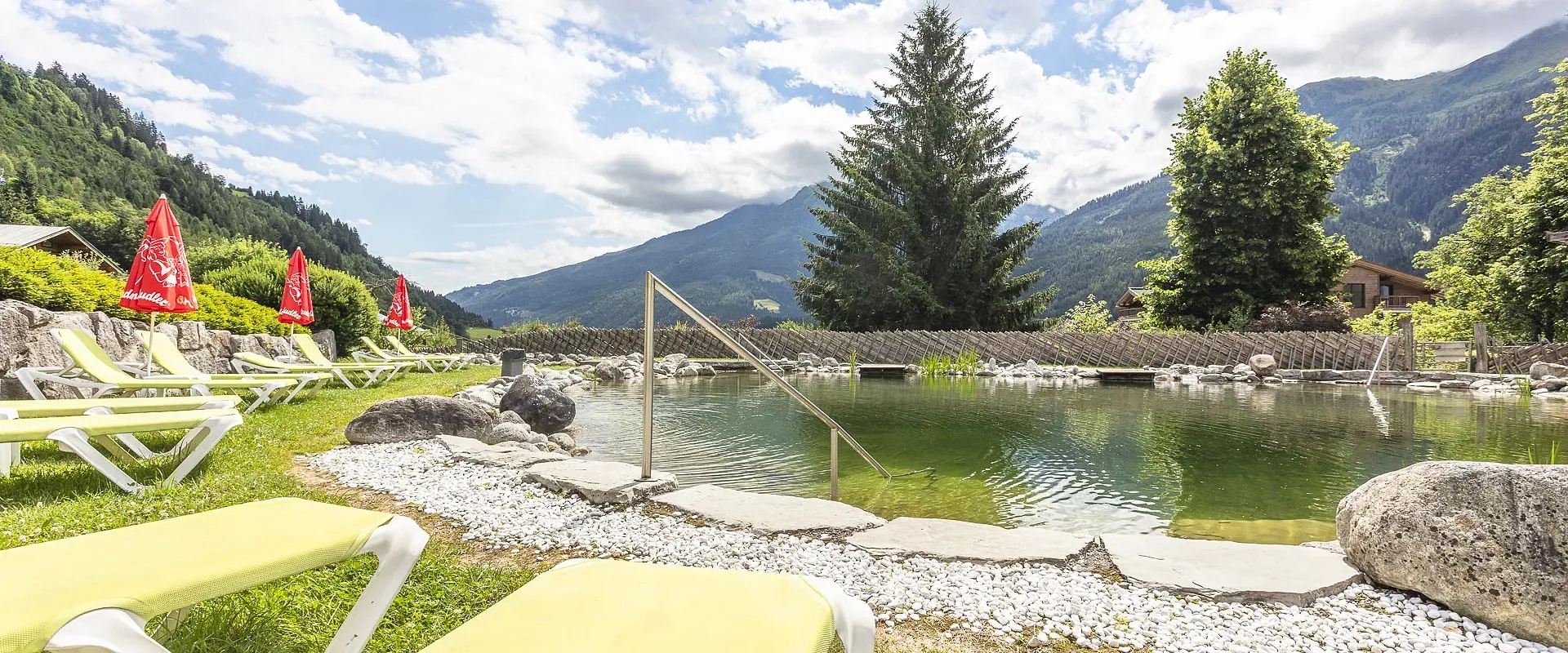 Ein idyllischer Teich mit Steinumrandung in einer Berglandschaft. Am Ufer stehen gelbe Liegestühle, umsäumt von Bäumen und unter einem klaren Himmel.