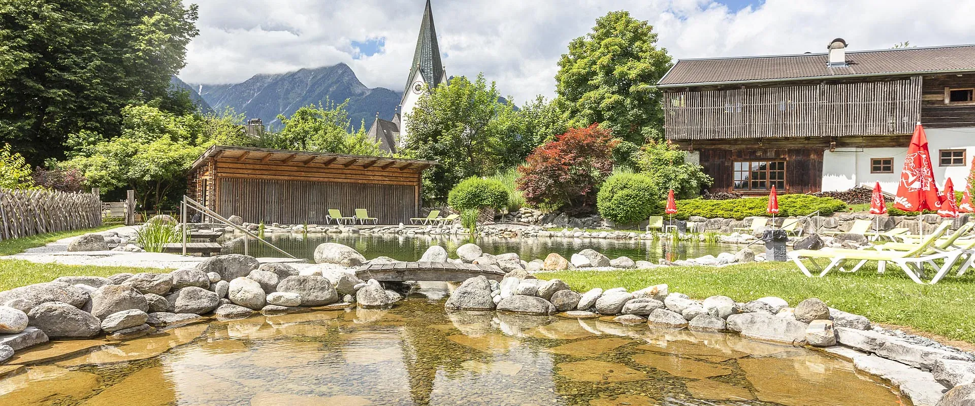 Ein idyllischer Garten mit einem Teich, umgeben von Steinen und Liegestühlen. Im Hintergrund sind ein altes Gebäude und Berge unter einem bewölkten Himmel zu sehen.