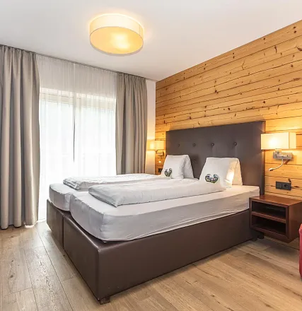 Gemütliches Schlafzimmer mit Holzwand, einem großen Fenster mit hellen Vorhängen und einem Doppelbett mit braunem Kopfteil und weißer Bettwäsche.