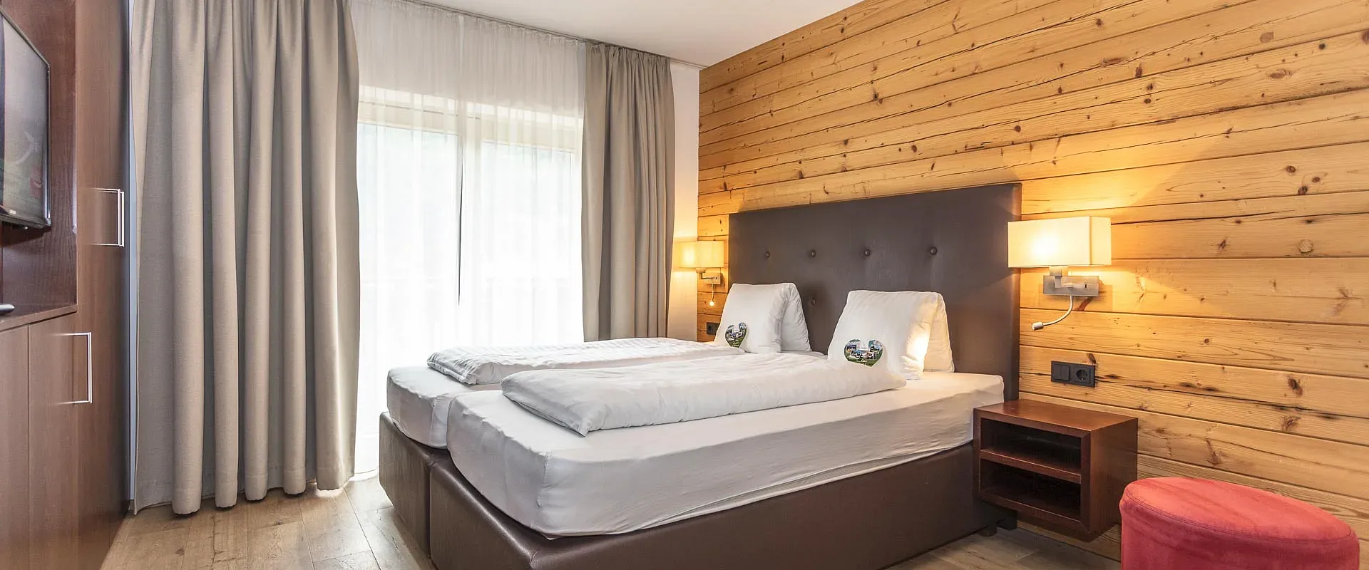 Gemütliches Hotelzimmer mit Holzwänden, einem Doppelbett mit weißer Bettwäsche, Nachttischlampen, einem roten Hocker und einem großen Fenster mit Vorhängen.