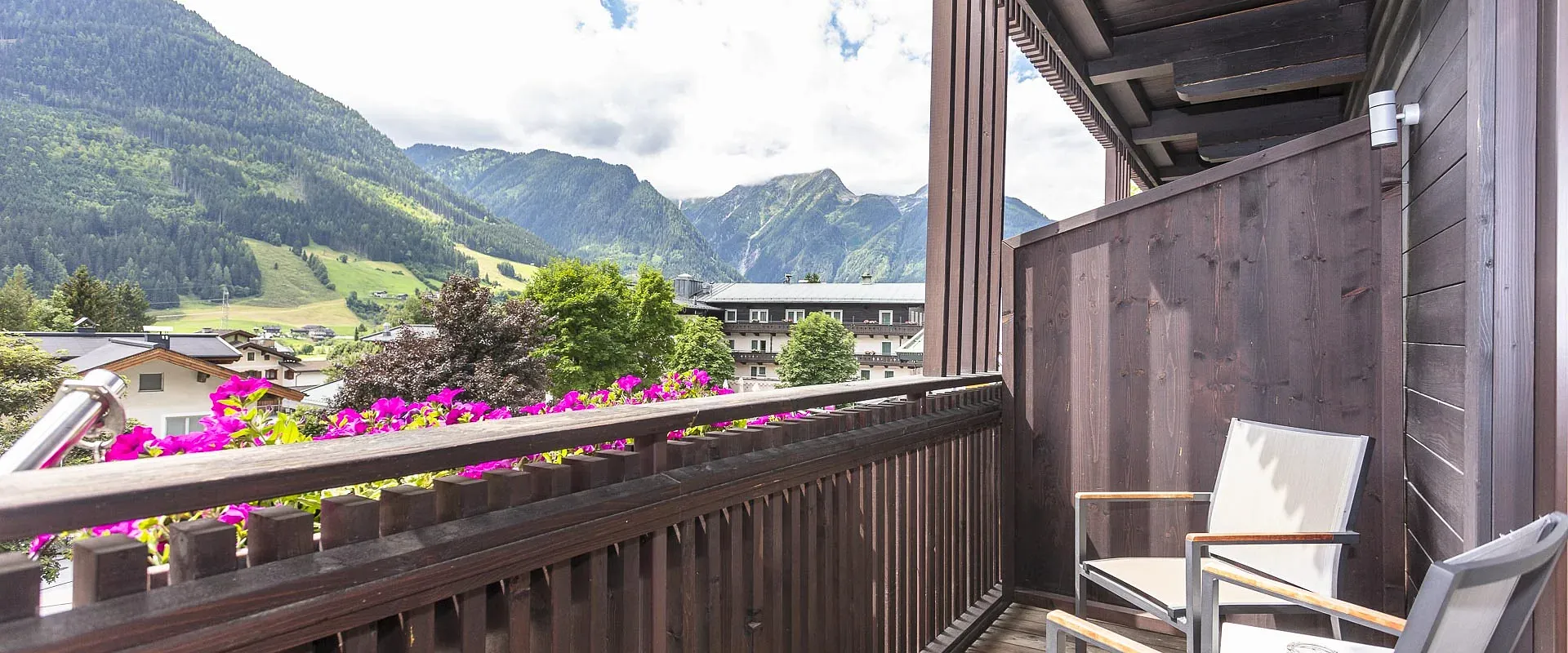 Ein hölzerner Balkon mit zwei Stühlen, umgeben von einer malerischen Berglandschaft. Farbige Blumen schmücken das Geländer unter einem blauen Himmel.