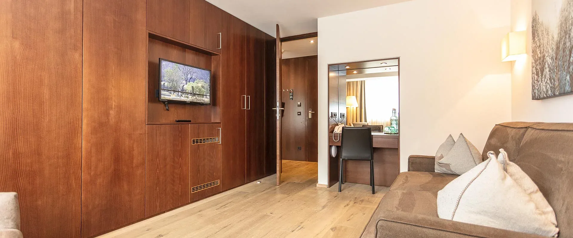 Helles, modernes Wohnzimmer mit Holzboden, beiger Couch und Holzwänden. Ein Flachbildfernseher ist an der Wand montiert. Ein Schreibtisch steht im Hintergrund.