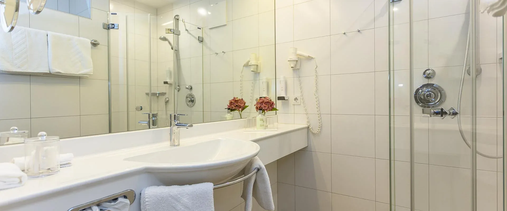 Modernes Badezimmer mit weißen Fliesen, großer Spiegelwand und Glasdusche. Auf der Arbeitsfläche stehen zwei dekorative Vasen mit roten Blumen.