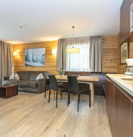 Gemütliches Wohnzimmer mit Holzwänden, einem Sofa, Esstisch und Stühlen. Ein großes Fenster mit Vorhängen lässt sanftes Licht herein, das Bild einer Winterlandschaft schmückt die Wand.