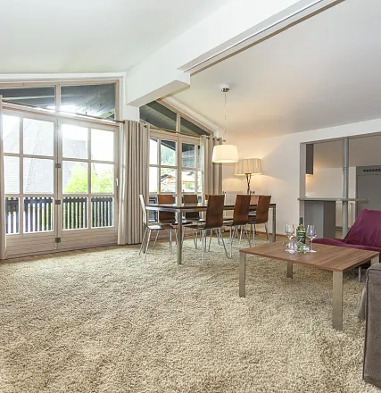 Helles Wohnzimmer mit großem Teppich, Holz-Esstisch und Stühlen. Große Fenster lassen viel Licht ein. Moderne Einrichtung und dezente Dekoration.