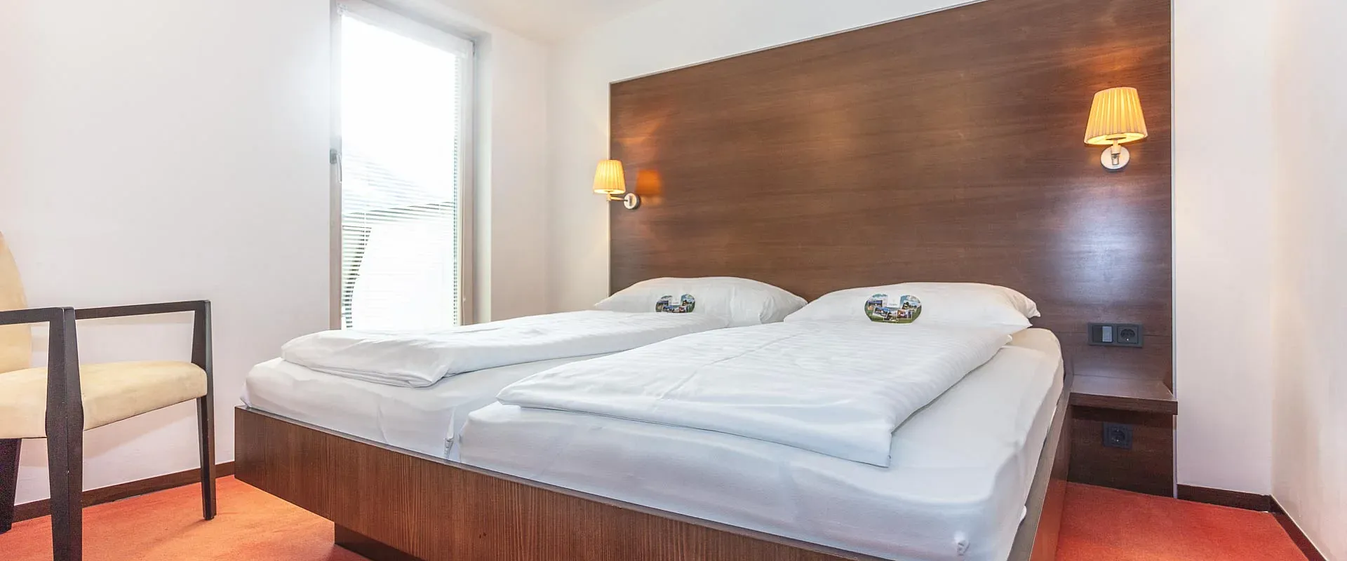 Schlichtes Hotelzimmer mit Doppelbett, weißer Bettwäsche und dunklem Holzkopfteil. Zwei Wandleuchten und ein Fenster sorgen für natürliches Licht.