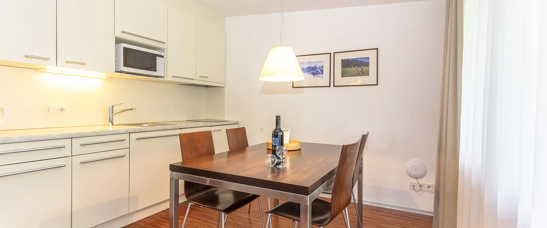 Moderne Küche mit Esstisch, vier Stühlen und schlichter Hängelampe. Helle Einbauküche, Holzfußboden und zwei Bilder an der strahlend weißen Wand.