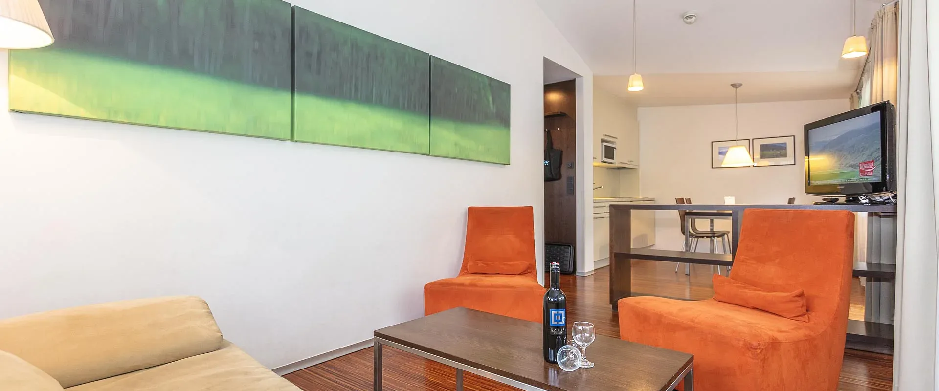 Modernes Wohnzimmer mit Holzboden, beigem Sofa, zwei orangefarbenen Sesseln und einem schmalen Couchtisch. An der Wand hängt ein abstraktes Bild.