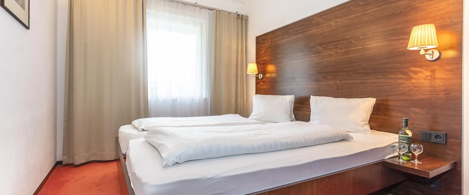 Gemütliches Hotelzimmer mit zwei Einzelbetten, einem großen Holzkopfteil, weißen Kissen und Bettdecken. Ein kleiner Tisch mit einer Flasche Wein steht daneben.