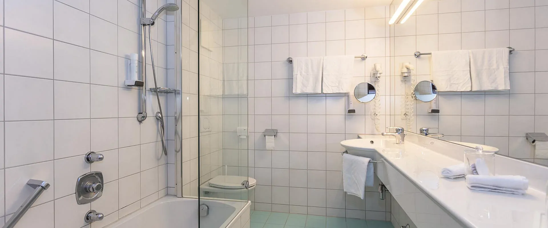 Modernes Badezimmer mit weiß gefliesten Wänden, einer Badewanne mit Duschkabine, einem großen Spiegel über dem Waschbecken und zwei Wandlampen.