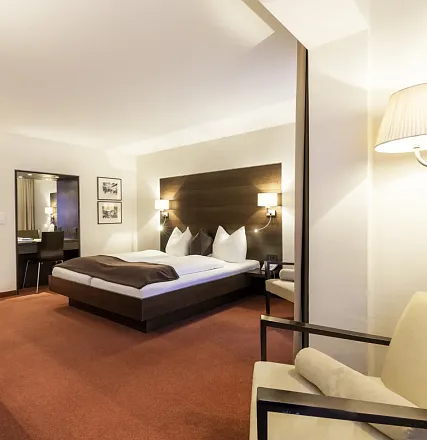 Modernes Hotelzimmer mit großem Bett, dunklem Holz-Kopfteil, zwei Nachttischlampen, Schreibtisch mit Stuhl und Wandkunst. Dezente Beleuchtung und gemütliche Atmosphäre.