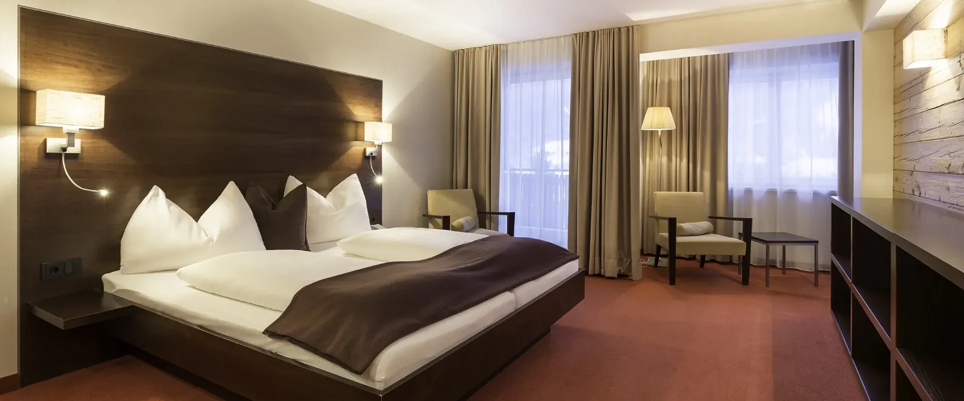 Elegantes Hotelzimmer mit Doppelbett, modernen Lampen, zwei Sesseln und einem großen Fenster mit Vorhängen. Der Raum ist in warmen Erdtönen gehalten.