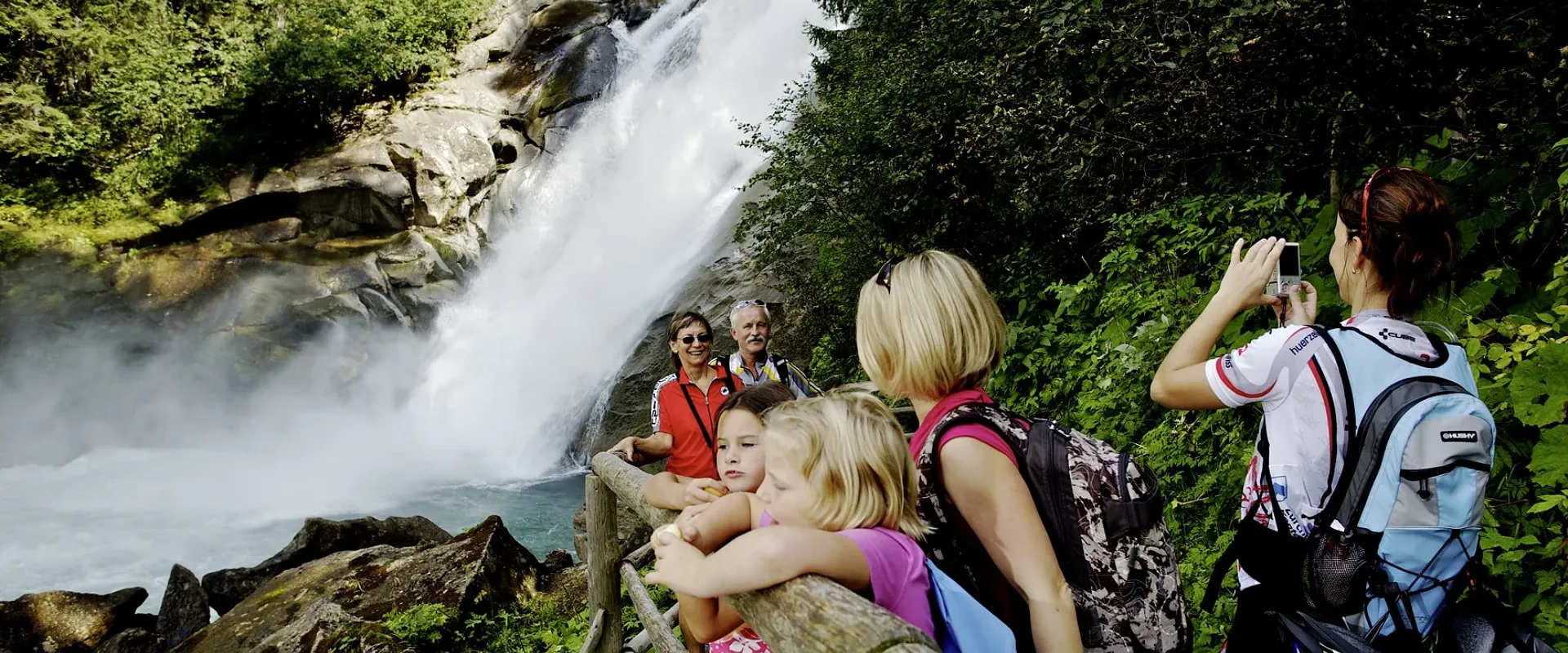 Eine Gruppe von Menschen steht vor einem beeindruckenden Wasserfall in einem Waldgebiet. Einige machen Fotos, während andere den Ausblick genießen.