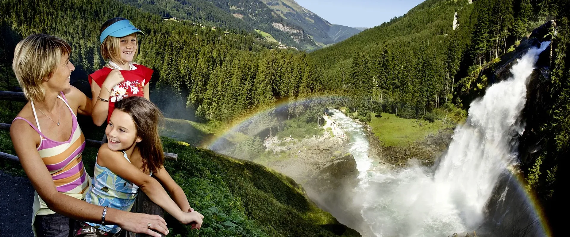 Familie genießt die Aussicht auf beeindruckenden Wasserfall inmitten grüner Berglandschaft mit Regenbogen. Klare blaue Himmel sorgt für sonnige Beleuchtung.