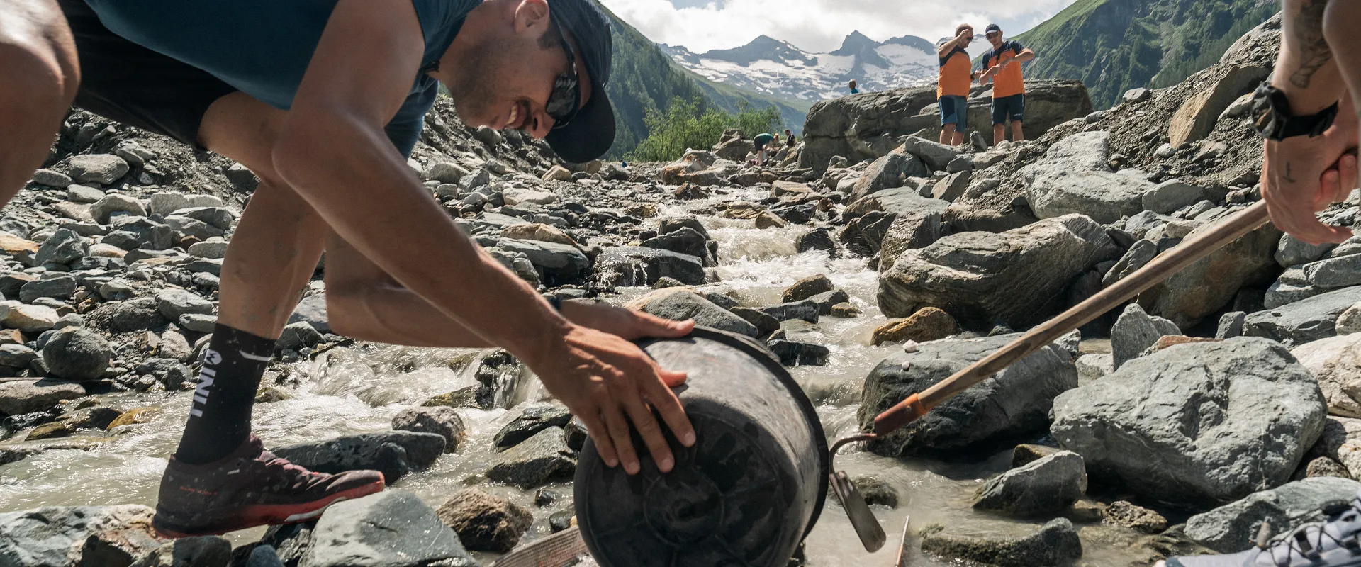 Personen in sportlicher Kleidung durchqueren einen flachen Gebirgsbach über Steine. Im Vordergrund wird ein großer Stein mit einem Hebel bewegt.