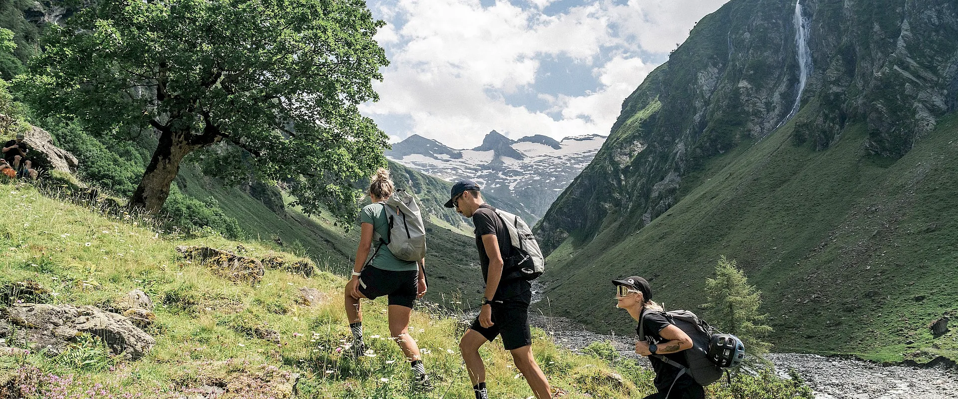 Drei Personen wandern in einer bergigen Landschaft mit grünen Hängen und einem blauen Himmel. Im Hintergrund sind schneebedeckte Gipfel zu sehen.