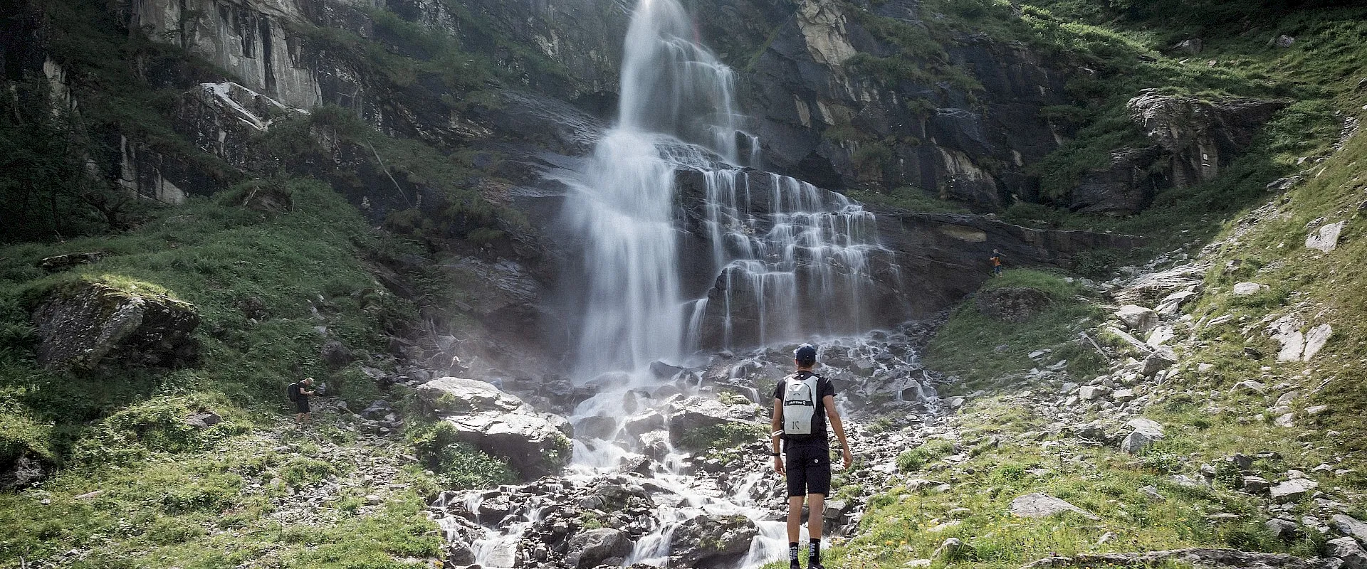 Person steht vor einem beeindruckenden Wasserfall in einer bergigen Landschaft mit üppiger Vegetation. Der Himmel ist teils bewölkt, die Szene wirkt ruhig.