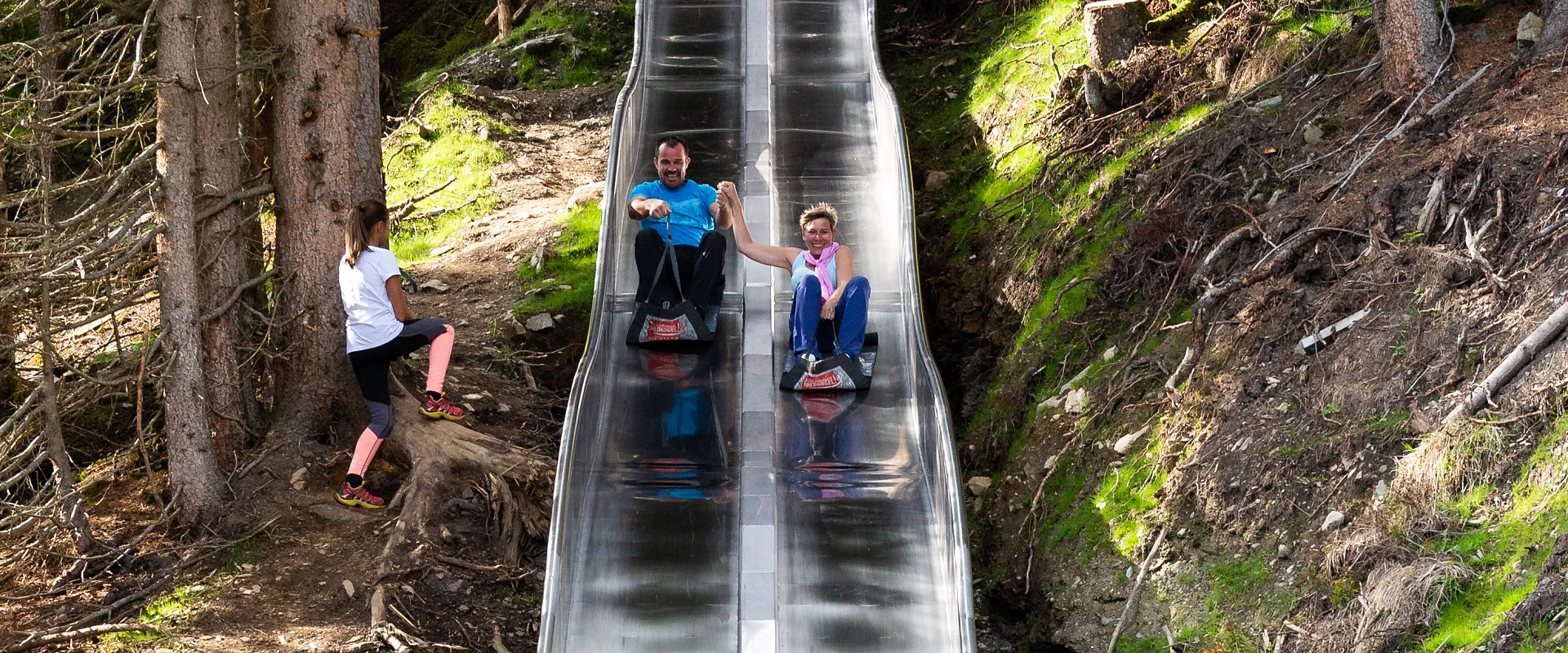 Zwei Personen rutschen fröhlich auf einer metallenen Rutsche im Wald hinunter, während eine dritte Person auf der linken Seite zusehen. Sonnenlicht scheint durch die Bäume.